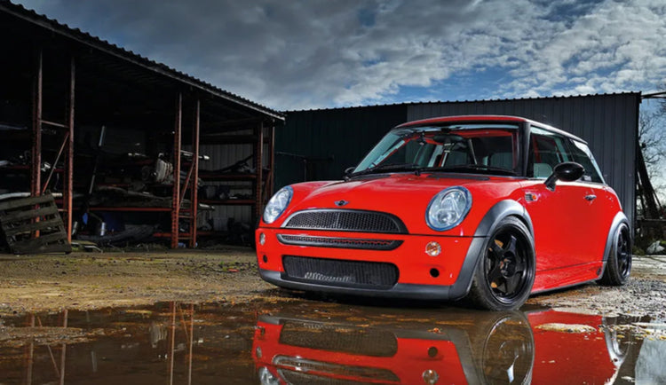 Red Mini Cooper car parked in front of a barn with a reflective surface below.